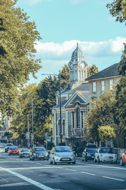 A view of a street in Childs Hill, featuring several parked cars along the pavement and a row of trees lining the sidewalk. In the background, there is a historic church with a clock tower, topped with a spire, and a decorative facade with arched windows and detailed brickwork. The street appears to be part of a residential area with a mix of modern and traditional buildings. The sky is partly cloudy with patches of blue, and the scene is well-lit by natural daylight. This setting exemplifies an urban environment suitable for home relocation and furniture transport services provided by Man with Van Childs Hill, with the focus on ongoing activities of moving and loading materials related to house removals.