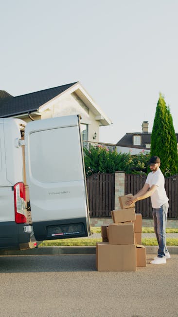 A man wearing a white t-shirt and jeans is standing outside a white moving van, unloading cardboard boxes during a home relocation. The van's rear door is open, revealing an empty interior, and the ma