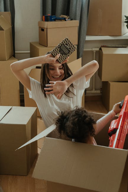 A family of three, including a young girl and her parents, are inside a well-lit room with large windows and light grey curtains, surrounded by numerous cardboard moving boxes of various sizes, some o