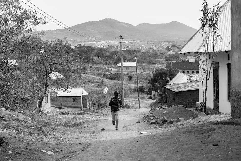 A young boy wearing a dark striped shirt and light-colored pants is walking along a dirt road in a rural residential area. The scene features uneven, rocky ground with some scattered debris and a small pile of soil on the right side. To the right, there is a partly constructed or weathered building with a metal roof, a window with a security grille, and a slim tree growing close to the wall. Further back on the right side, another small structure with a sloped roof is visible. On the left, there are trees and shrubs, with a few modest houses with metal or tiled roofs further down the road. Several wooden utility poles with overhead wires run along the street, connecting the area to the electrical grid. In the background, there are rolling hills or mountains under a partly cloudy sky, indicating a hilly or mountainous landscape. The image appears to be in black and white, emphasizing the textures and contrast of the rural environment. This scene could relate to house or village relocations, conveying a sense of moving logistics in a rural setting, as might be documented by [COMPANY_NAME] or similar services. 