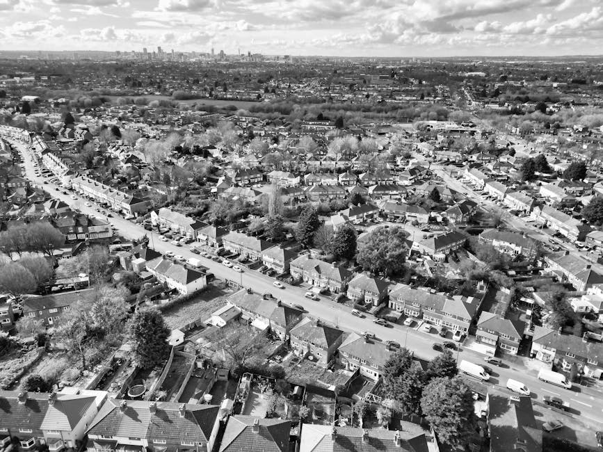 An aerial black-and-white photograph of a residential neighbourhood showing rows of terraced houses and semi-detached homes along winding streets, with trees and gardens interspersed throughout. The image captures a driveway with several cardboard boxes, piece of furniture wrapped in protective materials, and a large moving van parked on the pavement near a house entrance. Visible activity includes the loading process, with individuals handling and arranging packed items on trolleys and loading them into the van. The surrounding area extends towards the horizon, revealing an urban skyline with high-rise buildings in the distance. The scene illustrates elements of home relocation, including packing, furniture transport, and the logistical aspects of local house removals, with clear details of packaging materials such as cardboard boxes, protective blankets, and plastic wrap, alongside the moving vehicle ready for transit. Man with Van Childs Hill] is involved in the careful organization characteristic of professional moving services, aligning with the neighbourhood moving guide for Childs Hill Road to NW2.