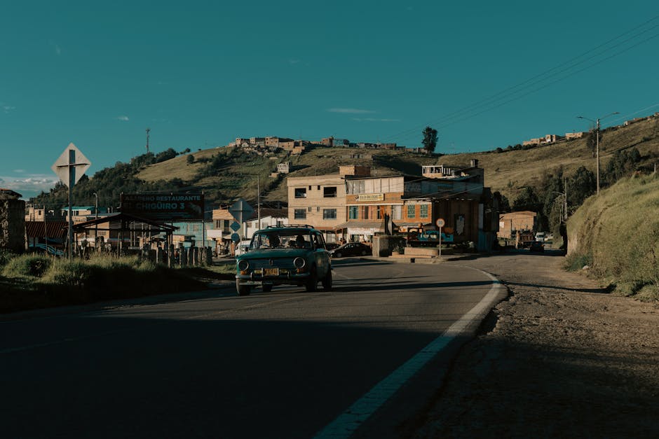 A young boy wearing a dark striped shirt and light-colored pants is walking along a dirt road in a rural residential area. The scene features uneven, rocky ground with some scattered debris and a small pile of soil on the right side. To the right, there is a partly constructed or weathered building with a metal roof, a window with a security grille, and a slim tree growing close to the wall. Further back on the right side, another small structure with a sloped roof is visible. On the left, there are trees and shrubs, with a few modest houses with metal or tiled roofs further down the road. Several wooden utility poles with overhead wires run along the street, connecting the area to the electrical grid. In the background, there are rolling hills or mountains under a partly cloudy sky, indicating a hilly or mountainous landscape. The image appears to be in black and white, emphasizing the textures and contrast of the rural environment. This scene could relate to house or village relocations, conveying a sense of moving logistics in a rural setting, as might be documented by [COMPANY_NAME] or similar services. 