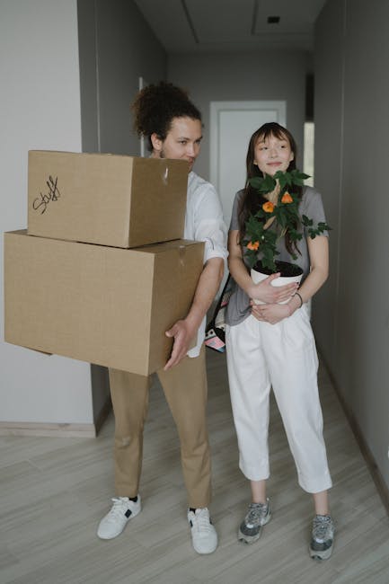 A woman and a young girl are inside a modern hallway preparing for a house relocation. The woman, dressed casually in beige trousers and a white shirt, is holding two large cardboard boxes stacked one on top of the other, both sealed with packing tape, and appears to be in the process of moving them. The girl, standing beside her, is holding a potted plant with green leaves and small orange flowers, wearing white pants, a grey T-shirt, and grey sneakers. The hallway has plain light-colored walls, a doorframe visible on the right, and a ceiling with built-in lighting. In the background, a glimpse of an adjacent room can be seen. This scene is part of a professional removals process involving packing and logistics, with [COMPANY_NAME] providing house moving services, and the environment suggests a careful and organized furniture transport and home packing activity.