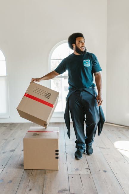 A man with dark skin and an afro hairstyle is inside a bright, minimally decorated room near a large arched window, holding a cardboard box wrapped with red tape in his right hand. He is dressed in a blue T-shirt and dark trousers, with a jacket tied around his waist. The room has light-colored walls, a wooden floor, and is well-lit by natural daylight streaming through the window. In front of him, on the floor, are two additional cardboard boxes, one stacked comfortably on top of the other, indicating a home relocation or furniture transport process. The images suggest an ongoing packing or loading activity in the context of house removals, aligned with the services offered by Man with Van Childs Hill for house and furniture moves in the Cricklewood and Childs Hill area.