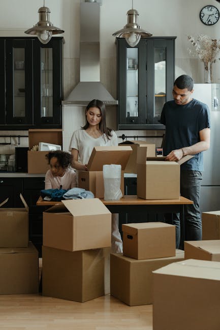 A woman and a young girl are inside a modern hallway preparing for a house relocation. The woman, dressed casually in beige trousers and a white shirt, is holding two large cardboard boxes stacked one on top of the other, both sealed with packing tape, and appears to be in the process of moving them. The girl, standing beside her, is holding a potted plant with green leaves and small orange flowers, wearing white pants, a grey T-shirt, and grey sneakers. The hallway has plain light-colored walls, a doorframe visible on the right, and a ceiling with built-in lighting. In the background, a glimpse of an adjacent room can be seen. This scene is part of a professional removals process involving packing and logistics, with [COMPANY_NAME] providing house moving services, and the environment suggests a careful and organized furniture transport and home packing activity.