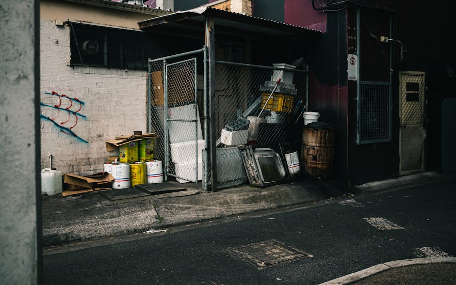 Outdoor provision of bulky waste items in Childs Hill, including stacked cardboard boxes, plastic containers and paint cans placed on the pavement near a covered storage area against a white brick wall. Inside the storage area, visible tools, a yellow plastic crate, and other miscellaneous materials are arranged on shelves and the ground. A large wooden barrel and white bowls are positioned close to the entrance of a dark building with a small window and metal door. The image captures a typical scene in a residential or commercial area where items may be prepared for disposal or collection, relevant to home relocation or furniture transport services provided by Man with Van Childs Hill as part of their removals operations. Clear lighting highlights the materials and surroundings, emphasizing the process of moving or clearing waste in the context of professional removals.