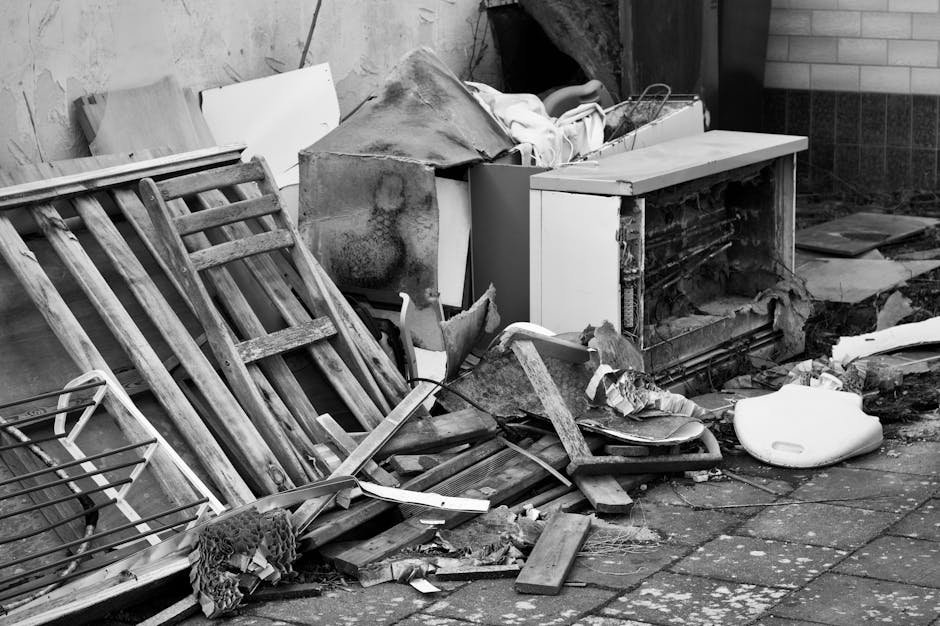 A black-and-white image showing a pile of discarded household items outside a property, including wooden furniture such as a disassembled bed frame or shelving unit leaning against a wall and a small cabinet with an open door. There are various debris scattered on the paved ground, like broken wooden planks, cardboard pieces, a piece of fabric or netting, a white plastic object, and some insulation or padding material. Some objects are partially covered with white fabric or protective wrapping. The scene appears to be in an outdoor area near the entrance or side of a building, with a brick wall visible on the right. The overall scene depicts items that are meant for removal, consistent with professional house removals or furniture transport services provided by Man with Van Childs Hill, highlighting the process of clearing out unwanted items during a home relocation or decluttering.