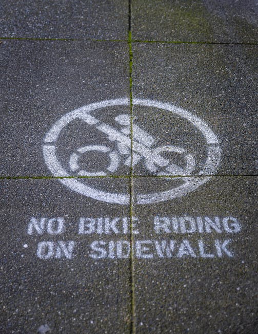 Close-up view of a concrete pavement with a white stencil marking illustrating a bicycle inside a circle, crossed out by a diagonal line, accompanied by the text 'NO BICYCLE RIDING ON SIDEWALK' below. The pavement consists of dark, textured concrete slabs with visible expansion joints and slight weathering. The scene is outdoors with natural lighting. This marking relates to public safety and pedestrian access regulations, relevant for home relocation and furniture transport activities near pavement areas, reflecting the importance of adhering to parking and loading rules in NW2, Chilms Hill, as outlined on the webpage for house removals and moving services by Man with Van Childs Hill.