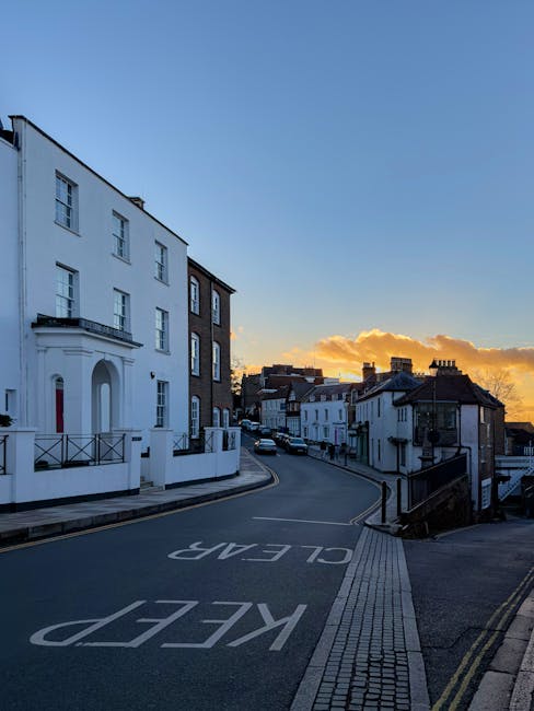 A wide view of a residential street in Chills Hill during sunset, showing a gentle curve with white and brick-built terraced houses lining both sides of the road. The pavement is clear, with a tactile paving strip along the edge for pedestrian accessibility. The sky is partly cloudy with orange and yellow hues from the setting sun, casting a warm glow over the scene. Parked cars are visible along the street, and the road features marked parking bays. The street appears quiet, with no visible moving vehicles or pedestrians. This setting exemplifies a typical UK neighbourhood where house removals and furniture transport might occur, especially considering the need to adhere to local loading and parking rules in NW2, as highlighted on the webpage for Man with Van Childs Hill.