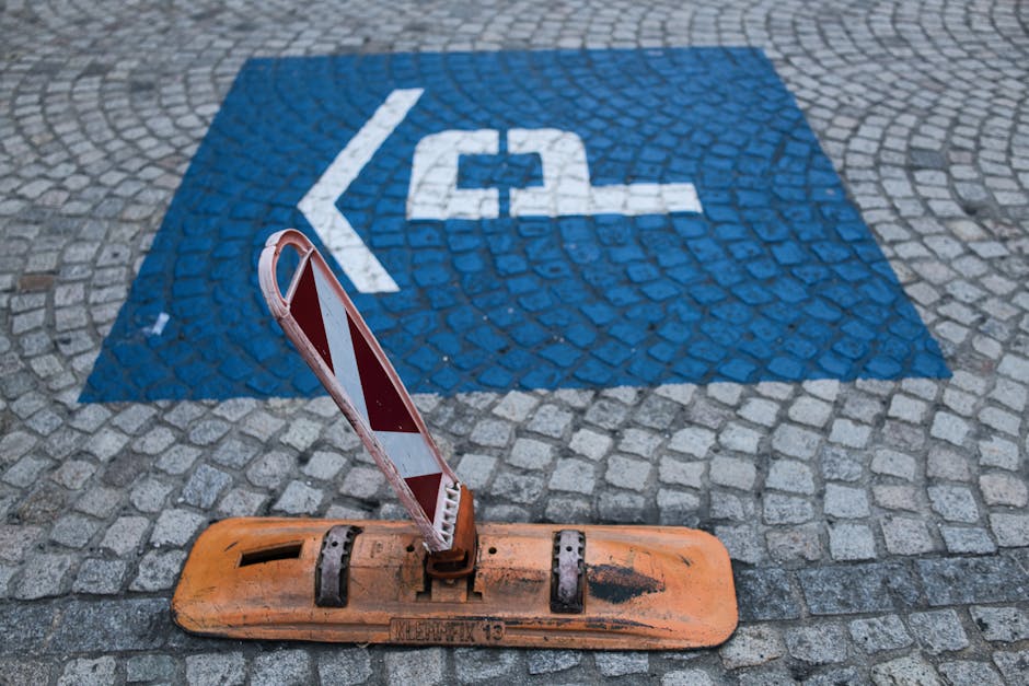 Close-up view of a concrete pavement with a white stencil marking illustrating a bicycle inside a circle, crossed out by a diagonal line, accompanied by the text 'NO BICYCLE RIDING ON SIDEWALK' below. The pavement consists of dark, textured concrete slabs with visible expansion joints and slight weathering. The scene is outdoors with natural lighting. This marking relates to public safety and pedestrian access regulations, relevant for home relocation and furniture transport activities near pavement areas, reflecting the importance of adhering to parking and loading rules in NW2, Chilms Hill, as outlined on the webpage for house removals and moving services by Man with Van Childs Hill.