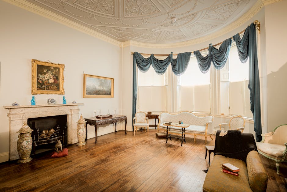 An interior view of a spacious, well-lit living room during a house removal process, with a high ornate ceiling and decorative plasterwork. The room features a large bay window dressed with dark blue, draped curtains arranged in swags, allowing natural light to fill the space. On the left side, there is a white marble fireplace with decorative vases and ornaments on the mantel, and a floral painting hung above it. In front of the fireplace, a small wooden side table with folding legs is positioned against the wall, holding a few small items. The room contains a set of vintage-style furniture: a long upholstered cream-colored sofa with gold trim, accompanied by matching armchairs, one of which is partially visible in the foreground with a black cloth draped over it. There are also wooden tables, one with a glass top, and a rectangular side table, all containing small objects. The floor is made of polished wooden planks, and there are no current items being moved or loaded, indicating that the furniture is still in place before packing or transporting. The overall scene reflects preparations within a period property managed by Man with Van Childs Hill as part of their professional removals and home relocation services. 