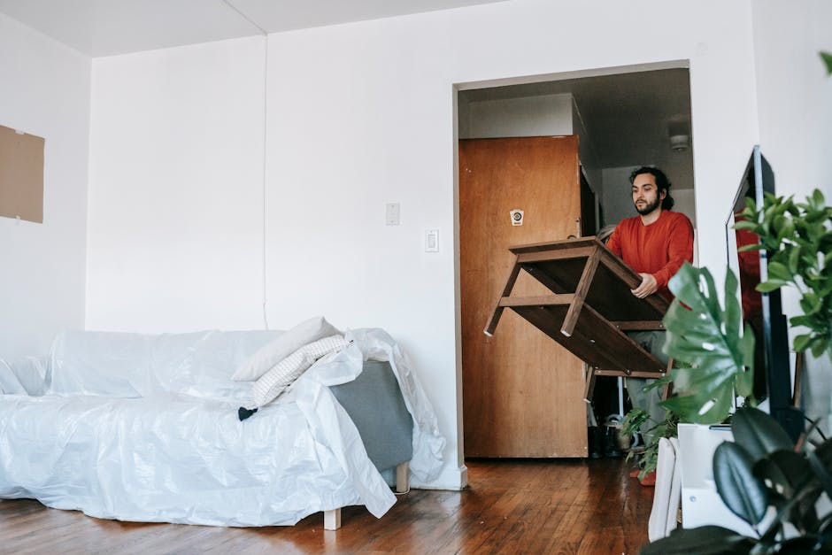 A man from Man with Van Childs Hill is seen inside a living room preparing for house relocation, standing behind a partially unfolded wooden dining table with a dark finish, holding one of its legs. The room features a white fabric-covered sofa with a pillow and some crumpled packing material on top, situated against a plain white wall. The flooring is wooden with a medium finish, and there is a doorway leading to a hallway or kitchen area in the background, partially obscured by the man and the table. To the right, a large potted plant with broad, dark green leaves is positioned near a television, which is turned off, along with some other empty or unpacked boxes indicating packing and moving activities. The environment appears well-lit with natural light, emphasizing the process of furniture transport and careful packing ahead of a home relocation handled by [COMPANY_NAME].