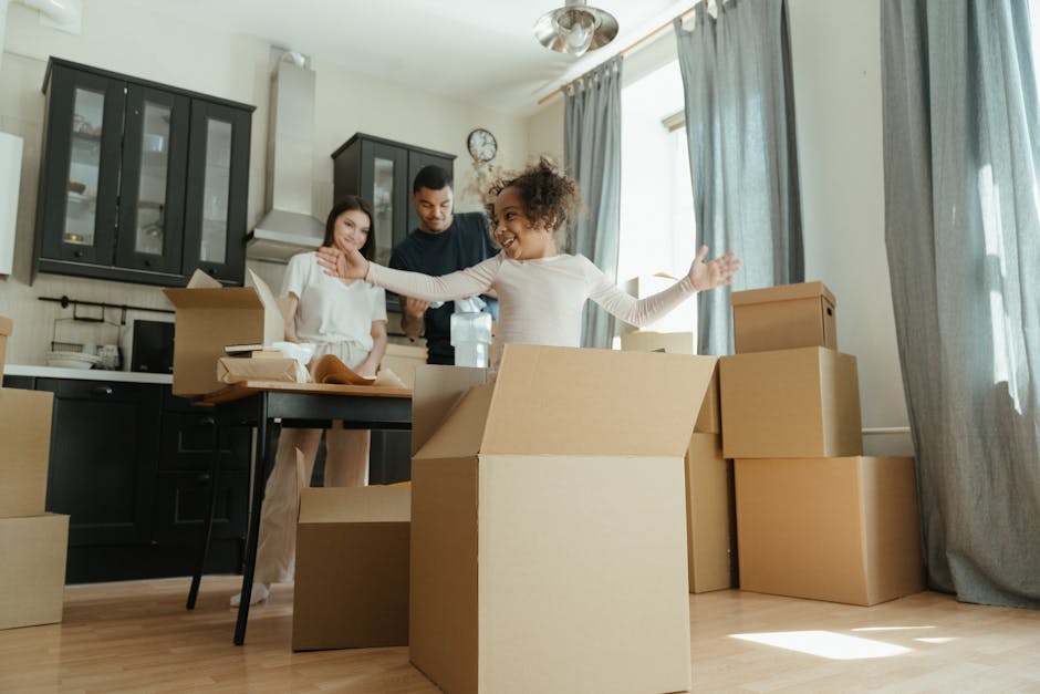 A family in a well-lit kitchen surrounded by numerous cardboard moving boxes of various sizes, some open and filled with packing materials, as they prepare for a home relocation. The family members are engaged in packing and unpacking activities; a young girl with curly hair, wearing a white top, is smiling and gesturing towards the boxes, while a woman and a man, both casually dressed, stand nearby assisting with packing. The kitchen features dark cabinets with glass doors, a white countertop, and has a modern, uncluttered appearance. The scene captures the process of furniture transport and packing as part of a house move managed by Man with Van Childs Hill, with natural light streaming in through large windows with light blue curtains, highlighting the ongoing loading process inside the property.