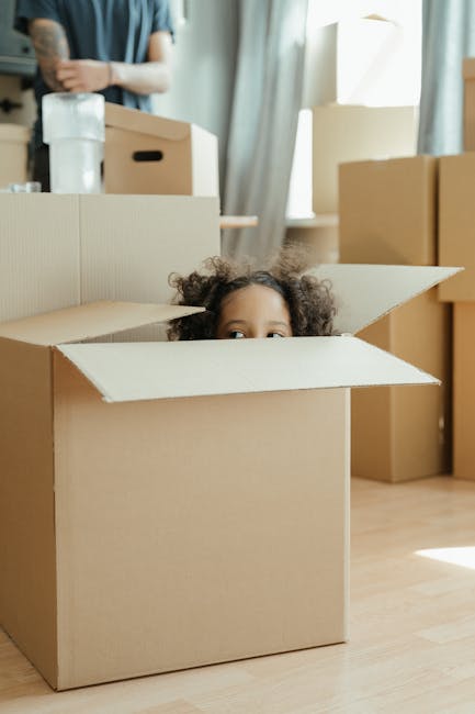 A young child with curly hair is sitting inside a partially open cardboard box placed on a wooden floor, surrounded by upright and stacked cardboard boxes in a well-lit room with large windows and sheer curtains. An adult, partially visible, is standing in the background near a countertop with packing materials, including a plastic bottle and more boxes, indicating a home relocation or packing process. The scene reflects furniture transportation, packing, and moving activities typical of a house removal, and the presence of various packed boxes suggests preparations for a moving service by Man with Van Childs Hill, supporting the theme of transparent moving prices in Childs Hill.