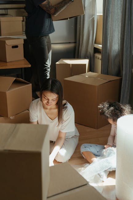 A family in a well-lit kitchen surrounded by numerous cardboard moving boxes of various sizes, some open and filled with packing materials, as they prepare for a home relocation. The family members are engaged in packing and unpacking activities; a young girl with curly hair, wearing a white top, is smiling and gesturing towards the boxes, while a woman and a man, both casually dressed, stand nearby assisting with packing. The kitchen features dark cabinets with glass doors, a white countertop, and has a modern, uncluttered appearance. The scene captures the process of furniture transport and packing as part of a house move managed by Man with Van Childs Hill, with natural light streaming in through large windows with light blue curtains, highlighting the ongoing loading process inside the property.