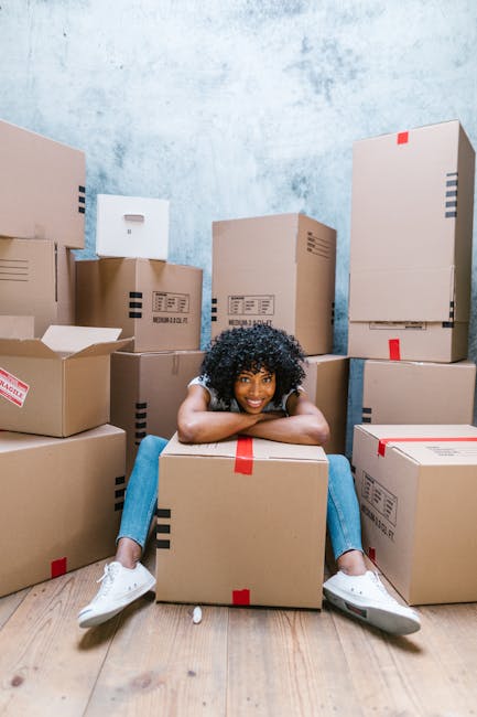 A young woman with curly black hair smiling while sitting cross-legged on a light wooden floor inside an empty room with a light blue textured wall. She is surrounded by numerous cardboard boxes of varying sizes, some sealed with red or black packing tape, and others partially open revealing packing materials. The boxes are stacked around her, with some on the floor and a few leaning against the wall. The woman is wearing casual clothing including a white top, blue jeans, and white sneakers. She appears to be in the process of moving home, possibly as part of a furniture transport or packing and moving service. The scene captures the preparation phase of a house relocation, with a focus on the stacked boxes and the indoor environment where home removals are being organized, as handled by companies like Man with Van Childs Hill.